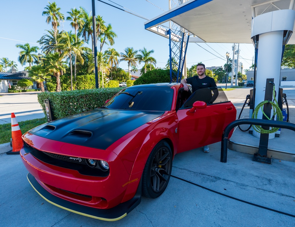 A man stands next to a red Dodge Challenger with the driver's door open at a gas station.
