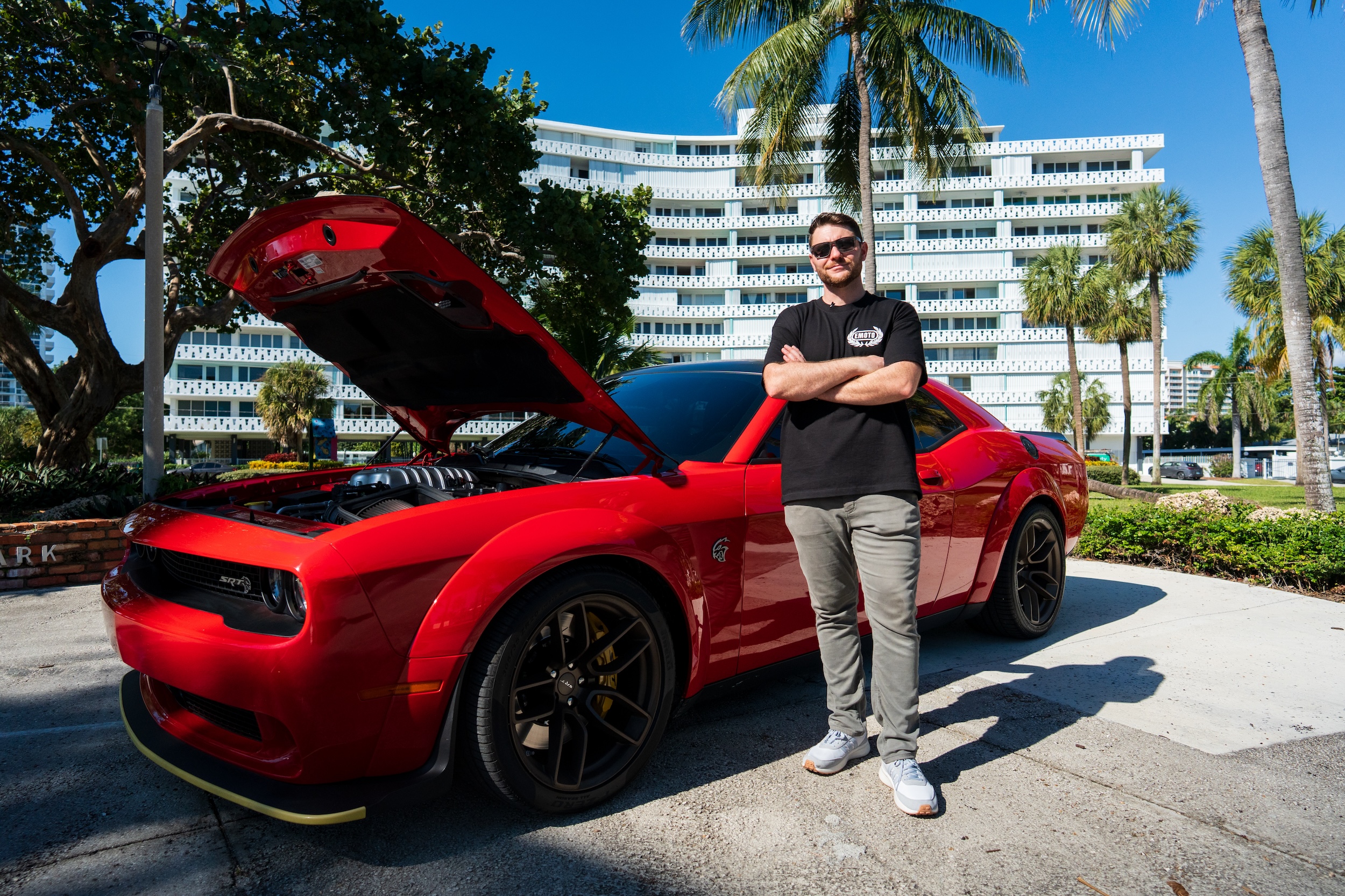 A man stands with arms crossed next to a red Dodge Challenger with its hood open outdoors.