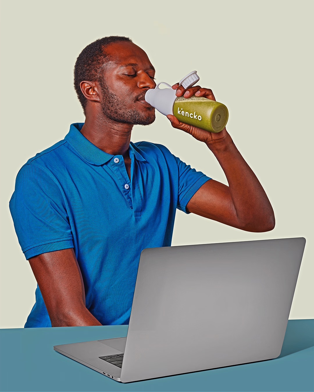 Person in blue shirt drinking from a bottle near a laptop.