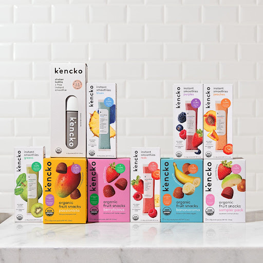 Assorted Kencko smoothie and snack boxes arranged on a marble surface against a white tiled background.