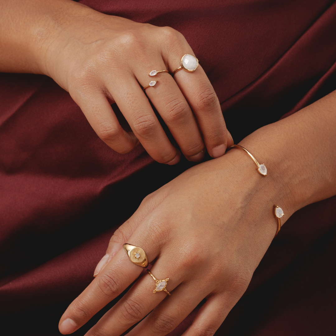Hands wearing gold rings and bracelets with gemstones on a burgundy fabric background.