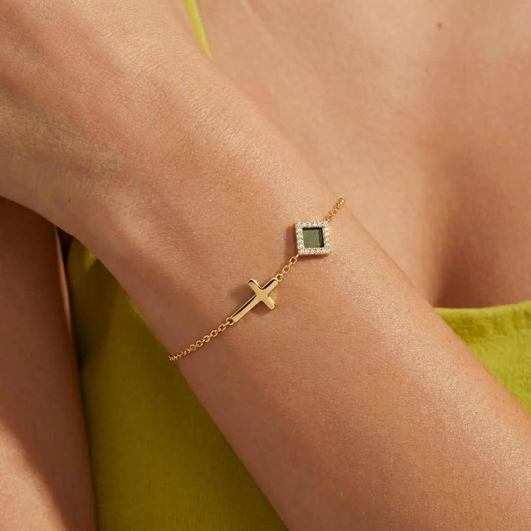 A close-up of a person's wrist wearing a delicate gold bracelet with a cross and green stone charm.
