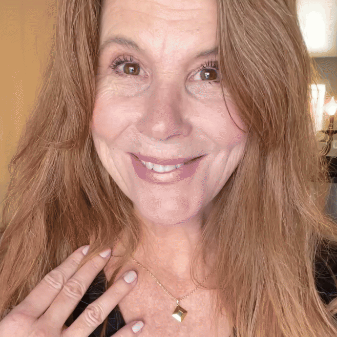 Smiling person with long hair wearing a necklace, touching their chest.