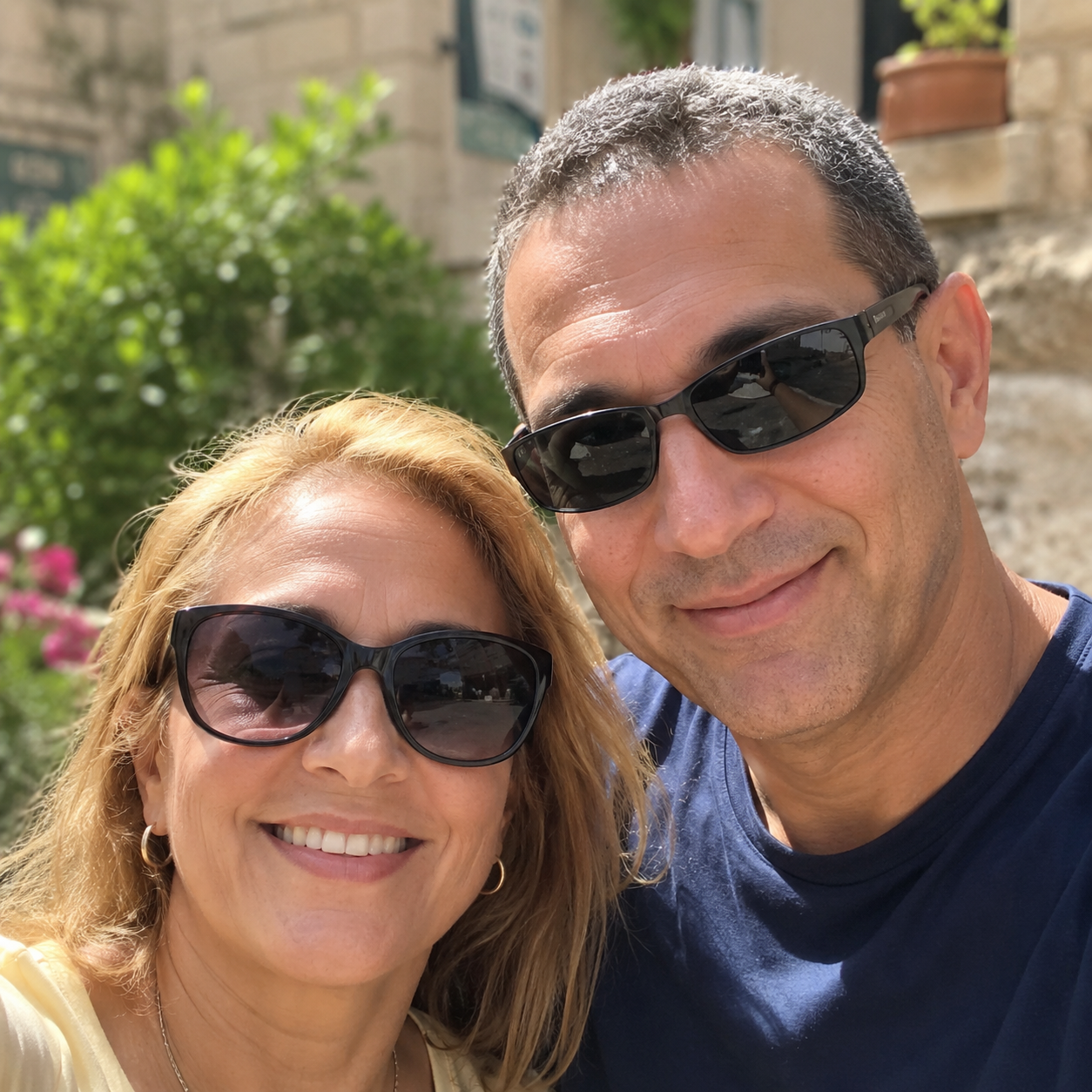 A smiling man and woman, both wearing sunglasses, pose for a selfie outdoors.