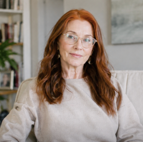 A woman with long red hair and glasses, wearing a sweater, smiles while sitting on a couch.