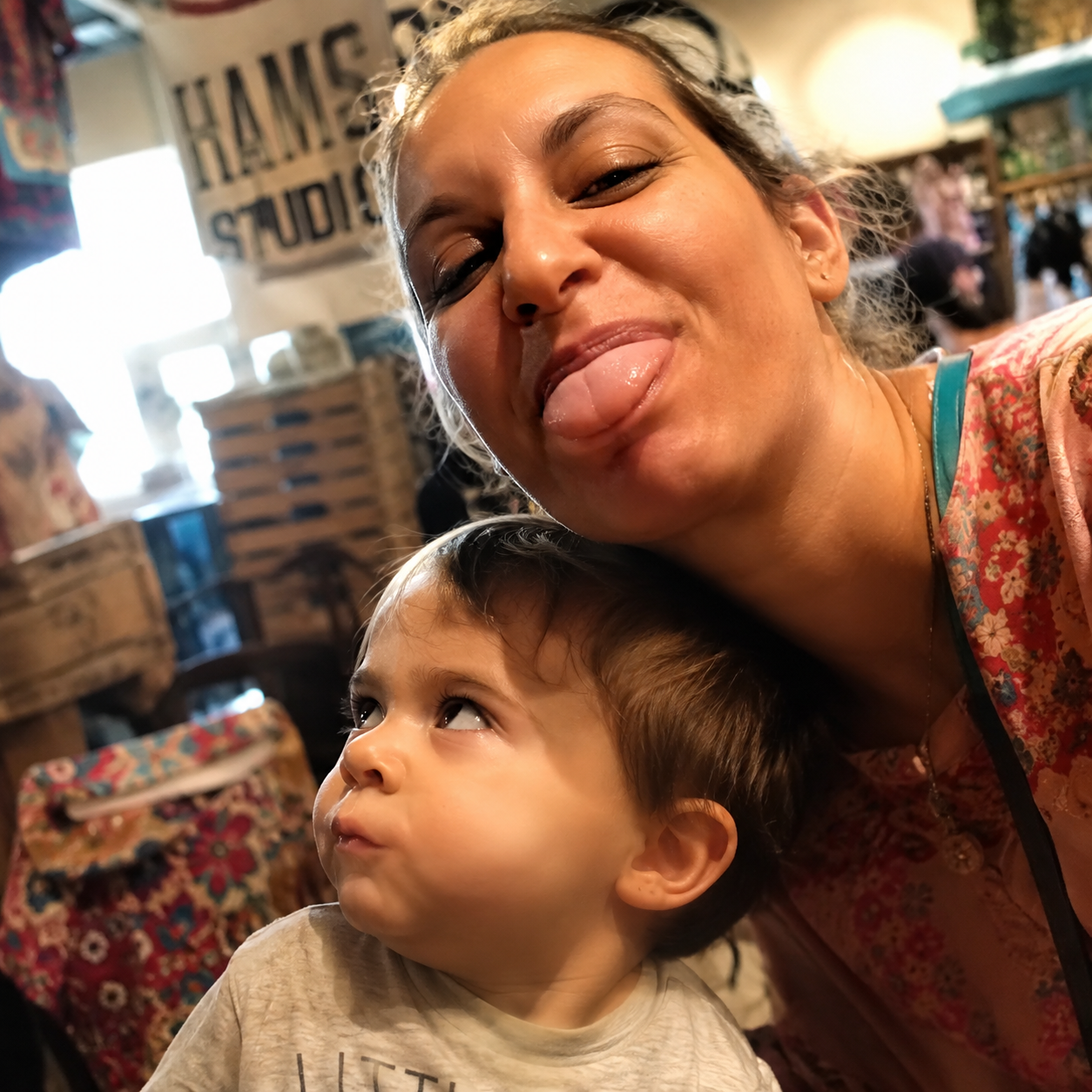 A woman playfully sticks her tongue out while taking a selfie with a young child looking upwards.