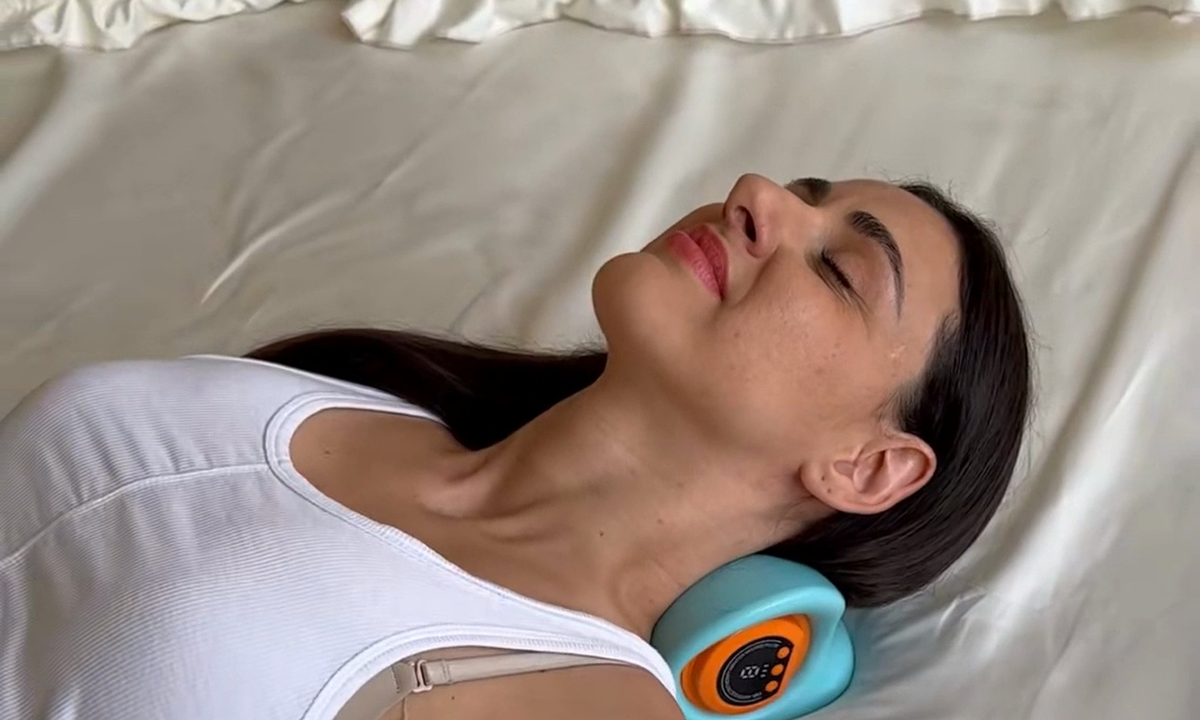 A woman with her eyes closed lies on her back using a blue electronic neck stretching device.