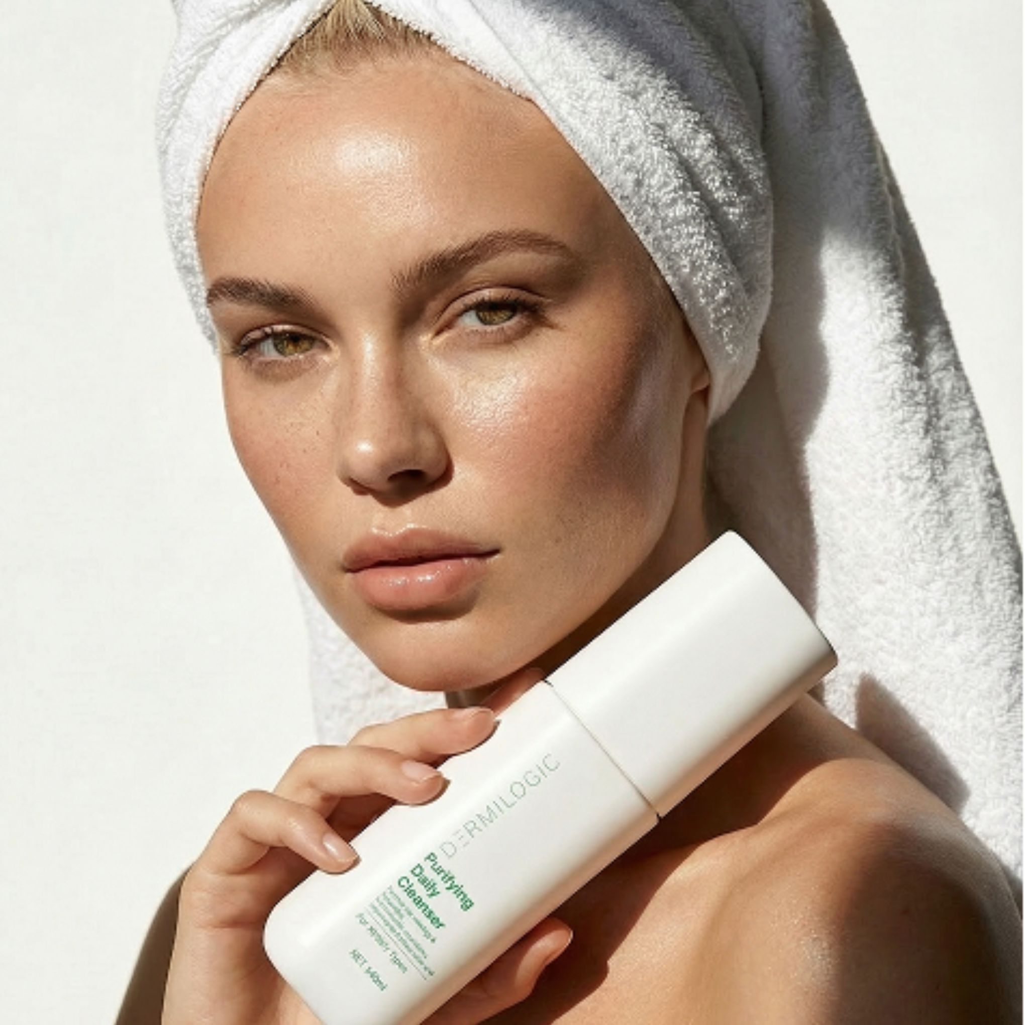 A woman with her hair wrapped in a white towel holds a bottle of Dermilogic facial cleanser.