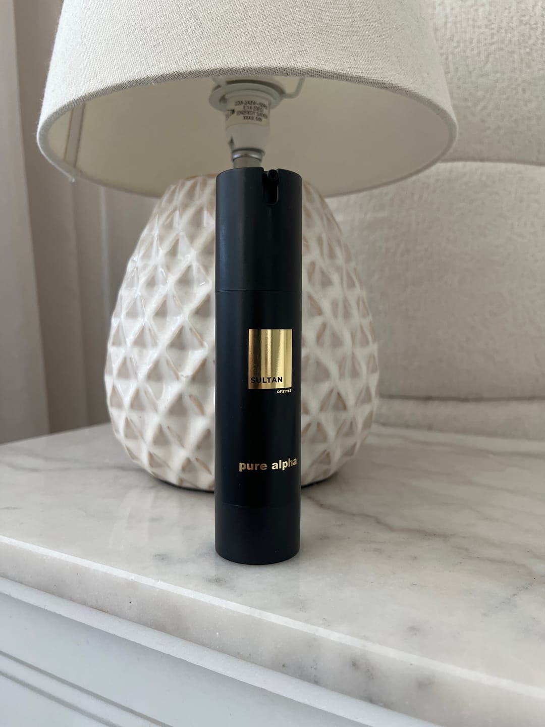 A black pump bottle of Sultan pure alpha sits on a marble surface in front of a textured white lamp.