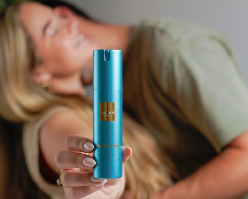 A woman's hand holds up a blue pump bottle with a gold label, with a couple blurred behind it.