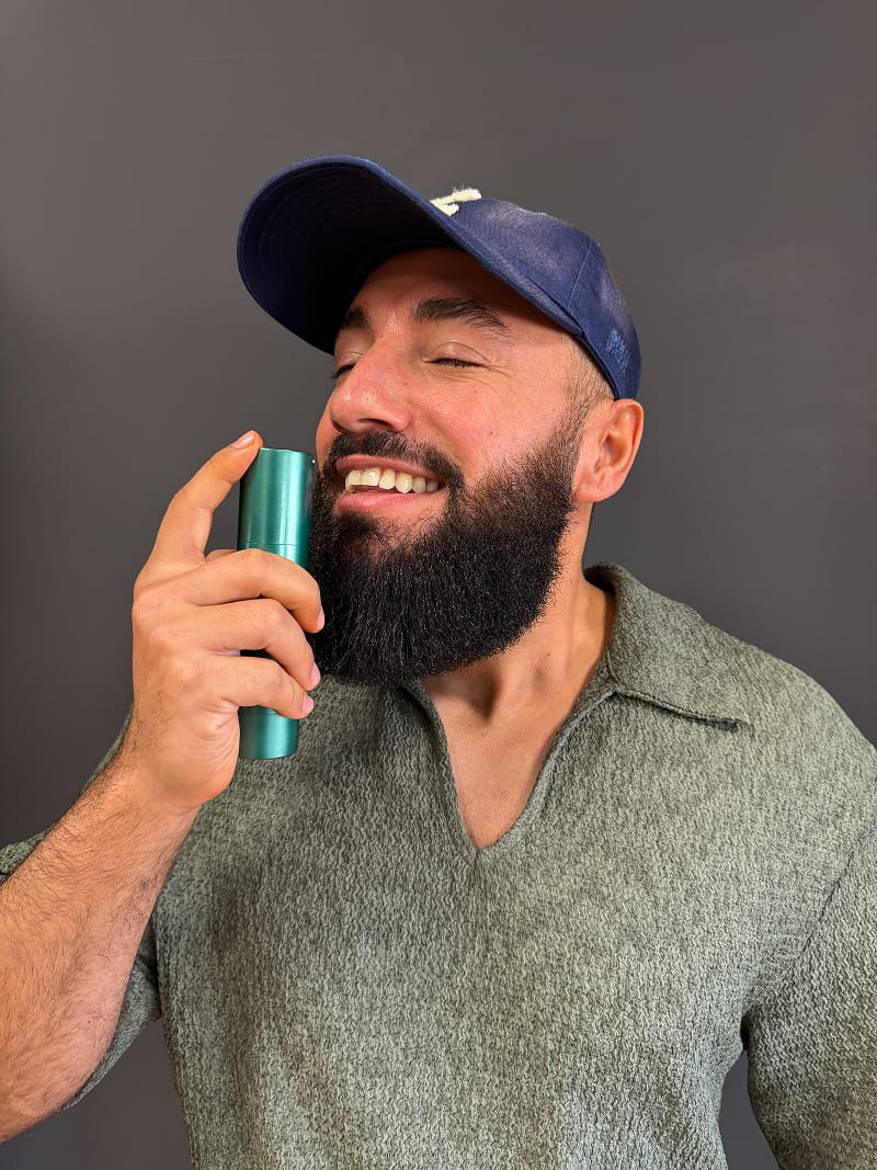 A smiling, bearded man in a blue cap holds a small teal bottle to his nose.