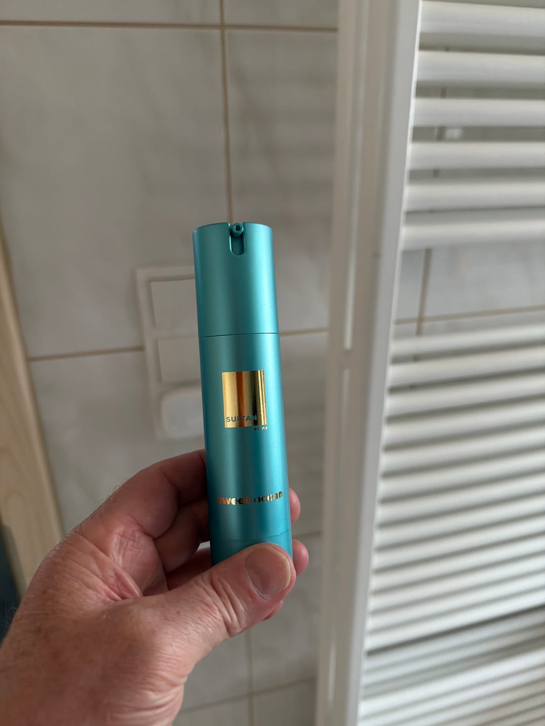 A hand holds a teal-colored pump bottle with a gold label against a white tiled wall.