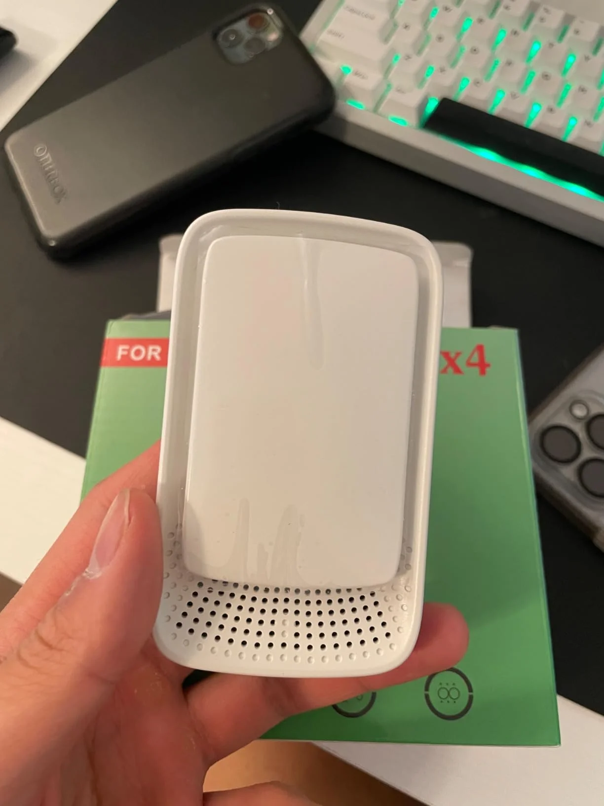 Hand holding a white electronic device with perforations, near a keyboard and phone.