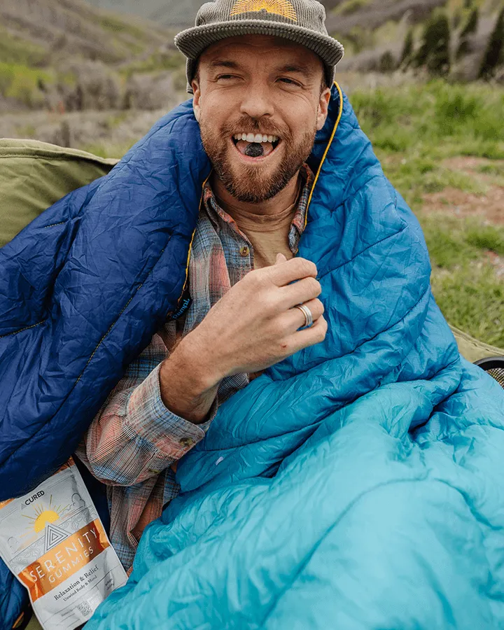 Man in a blue sleeping bag outdoors, holding a gummy.