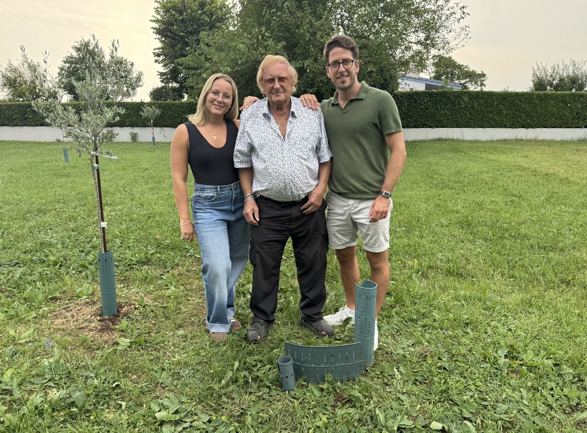 Three people standing on grass with small trees nearby.