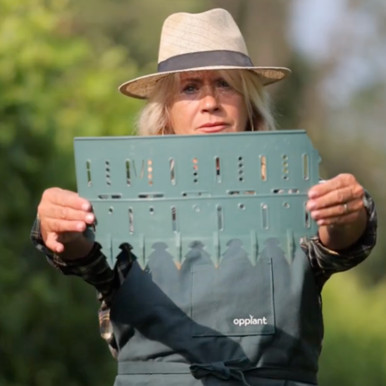 A person in a hat and apron holds up a green plastic gardening tool.