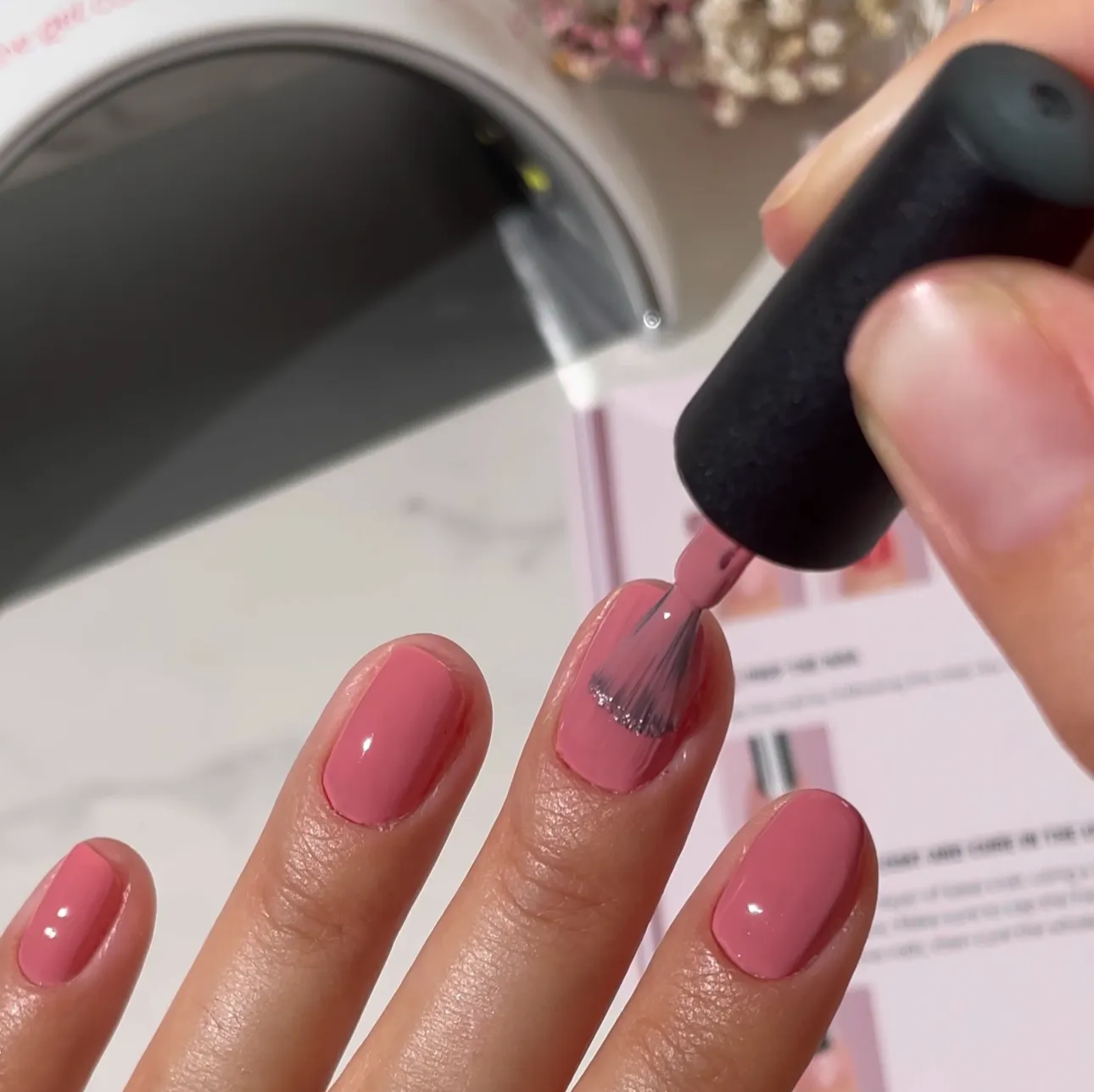 A close-up shot of a person applying a coat of shiny pink nail polish to their fingernails.