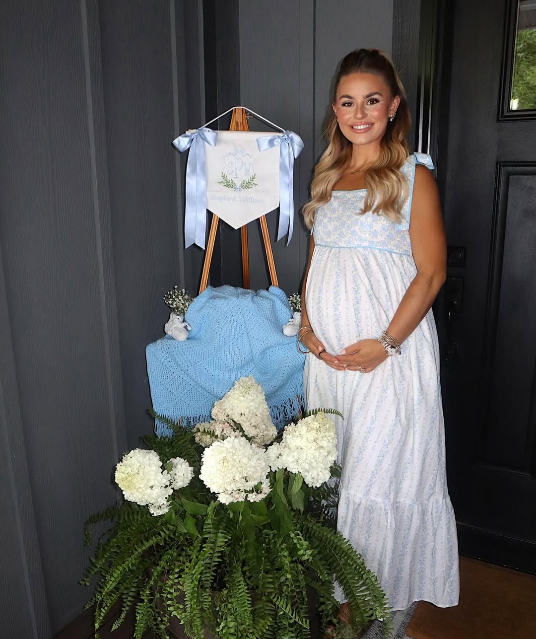 Pregnant woman in a blue dress stands by a decorated easel and potted plants.