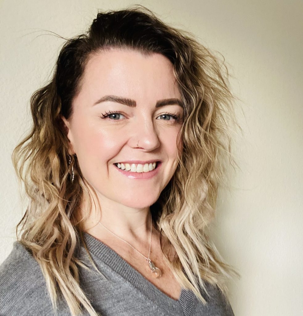 Smiling person with wavy hair and a grey top against a plain background.