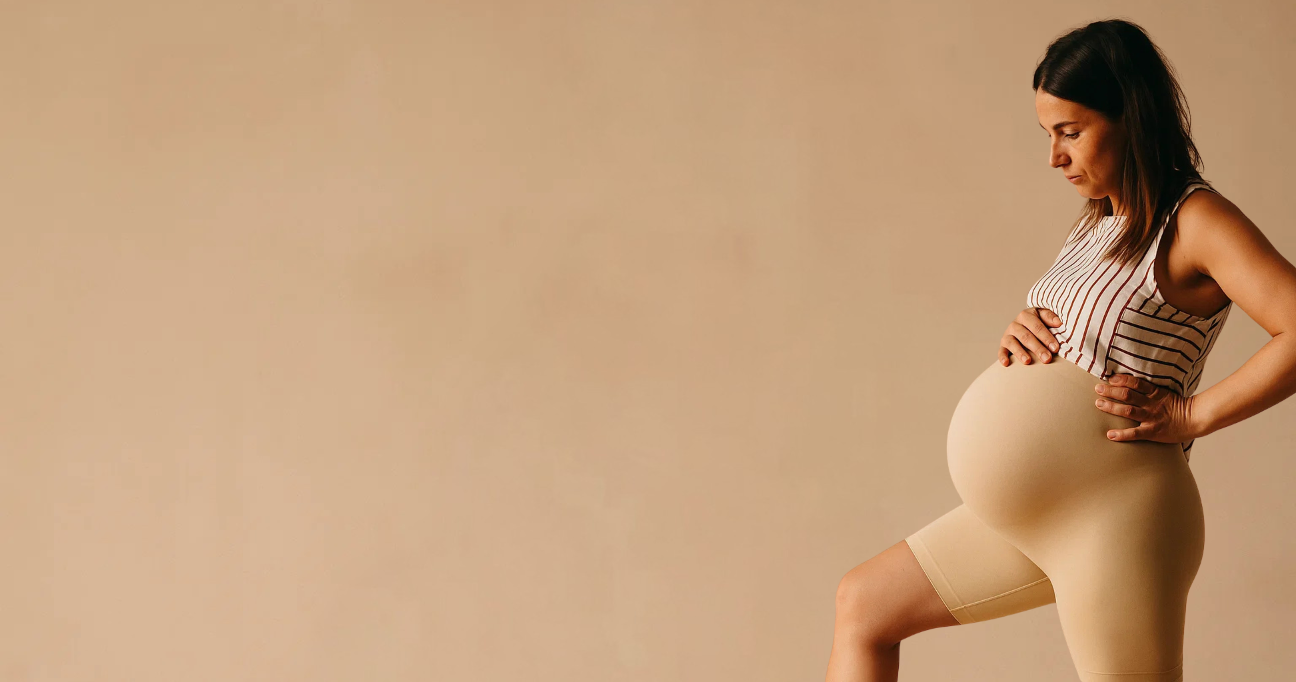 Pregnant woman in striped top and beige shorts, standing indoors.