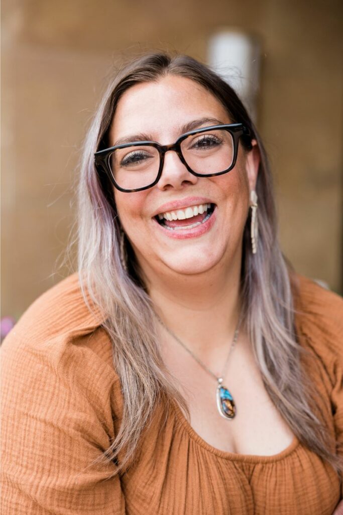 Smiling person with glasses and long hair, wearing a brown top and necklace.