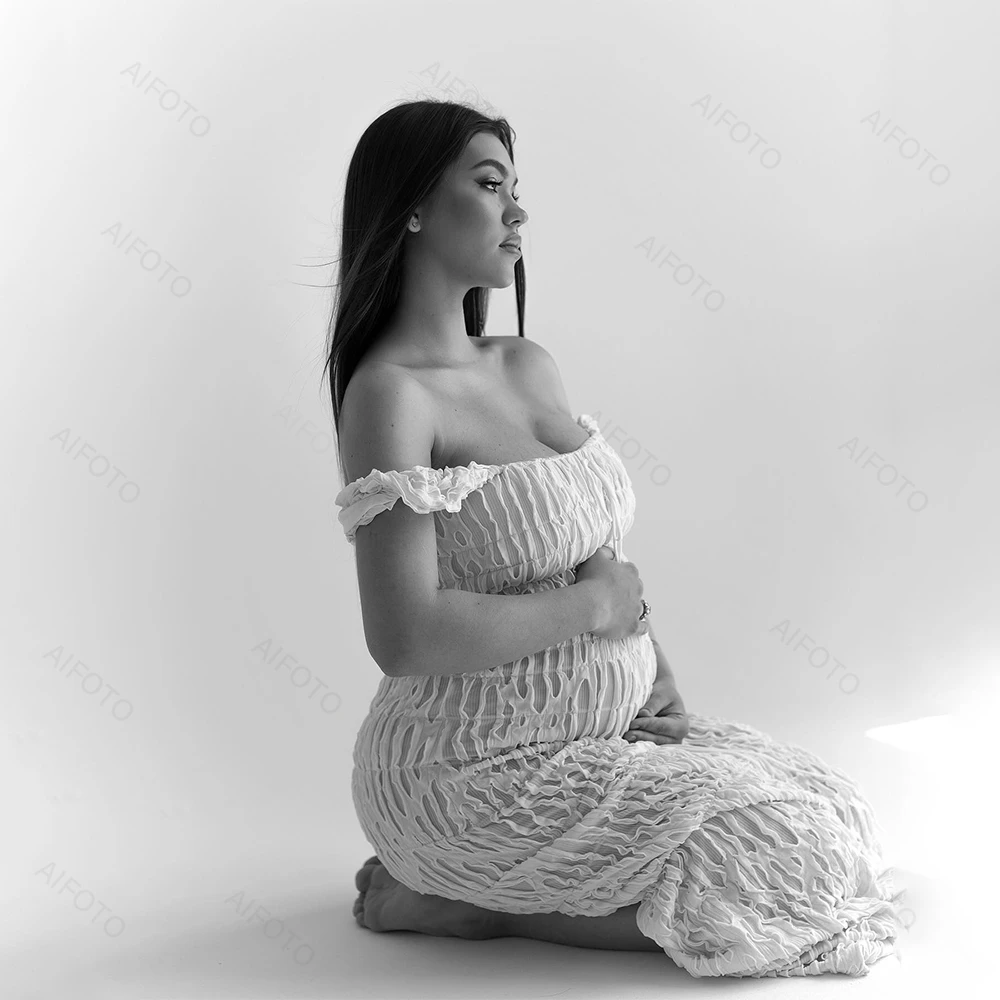 A pregnant woman in an off-the-shoulder dress kneels in profile, holding her belly against a white background.