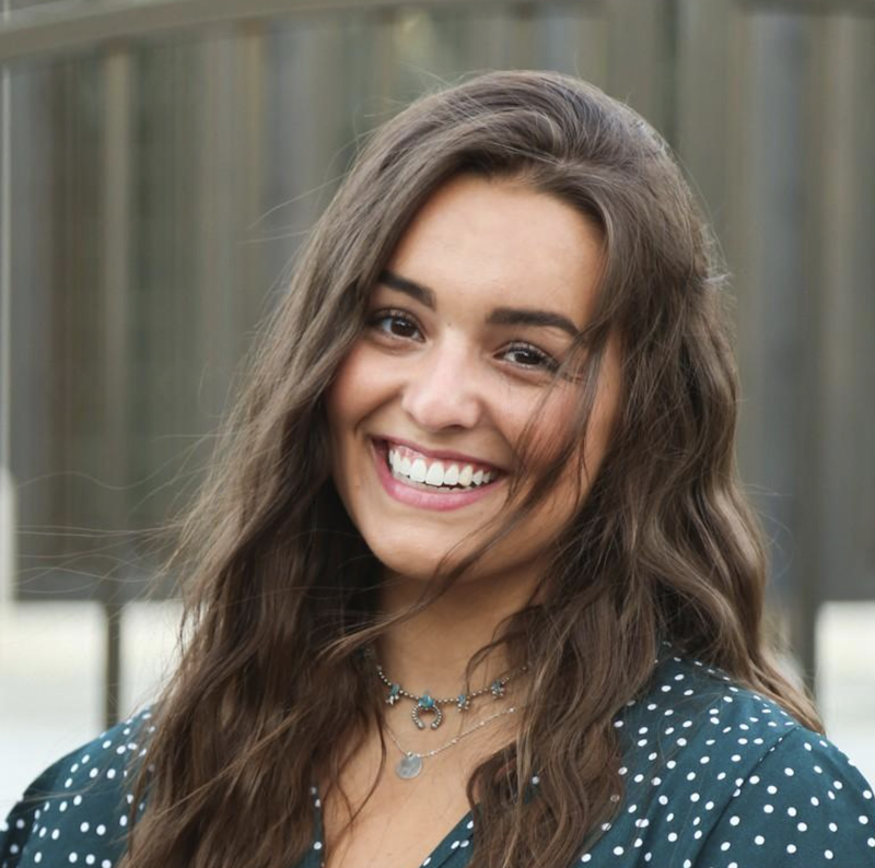 Smiling person with long hair wearing a polka dot shirt and necklace.
