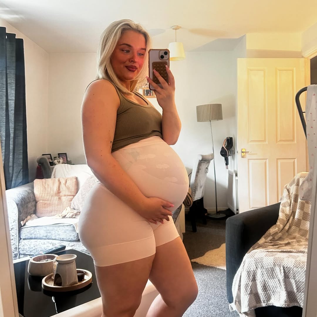 A person takes a mirror selfie in a living room, showcasing their pregnancy.