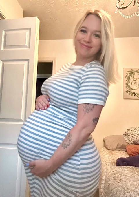 Pregnant woman in striped dress smiling and holding her belly in a bedroom.