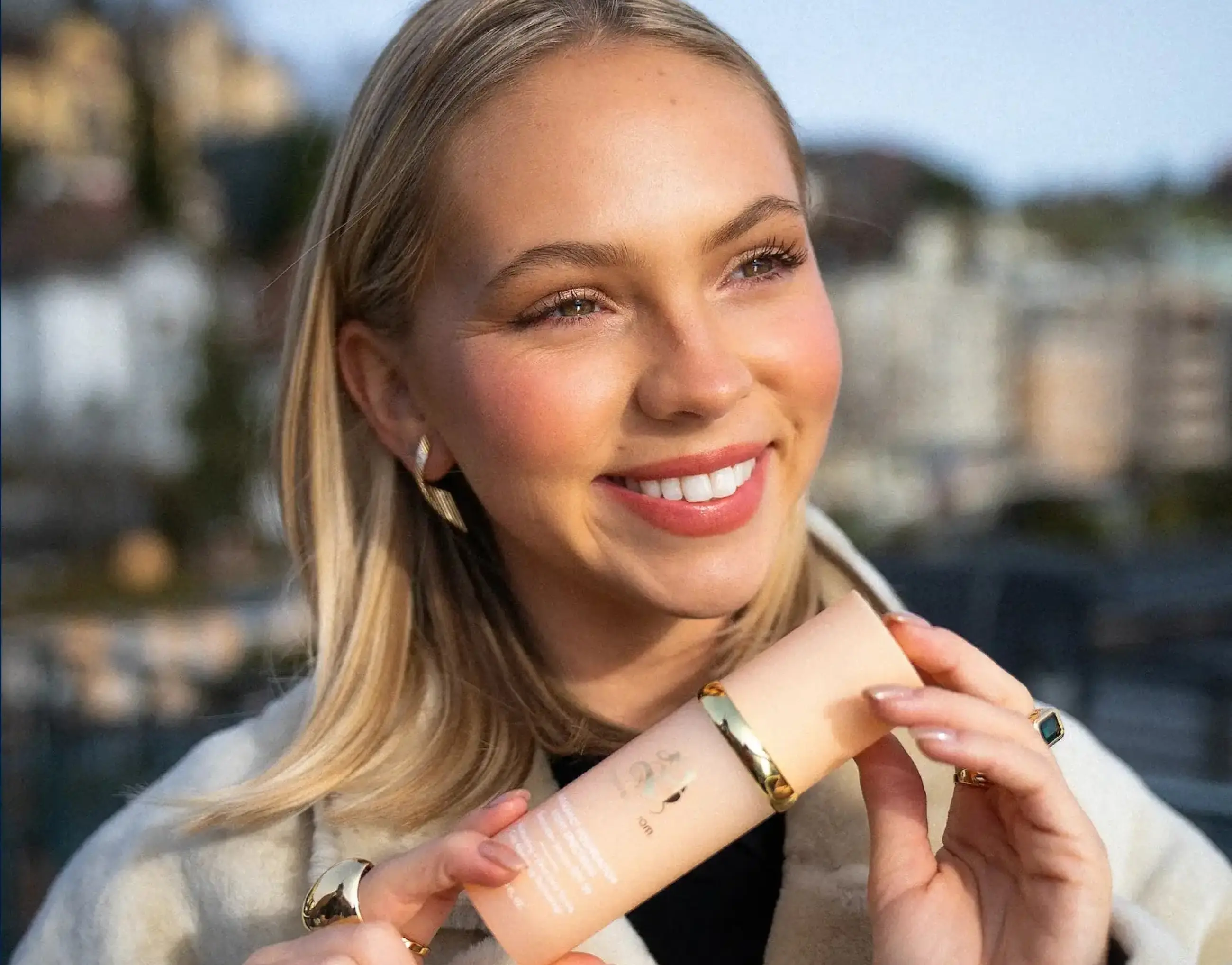 A smiling blonde woman outdoors holds a peach-colored bottle with a gold band.