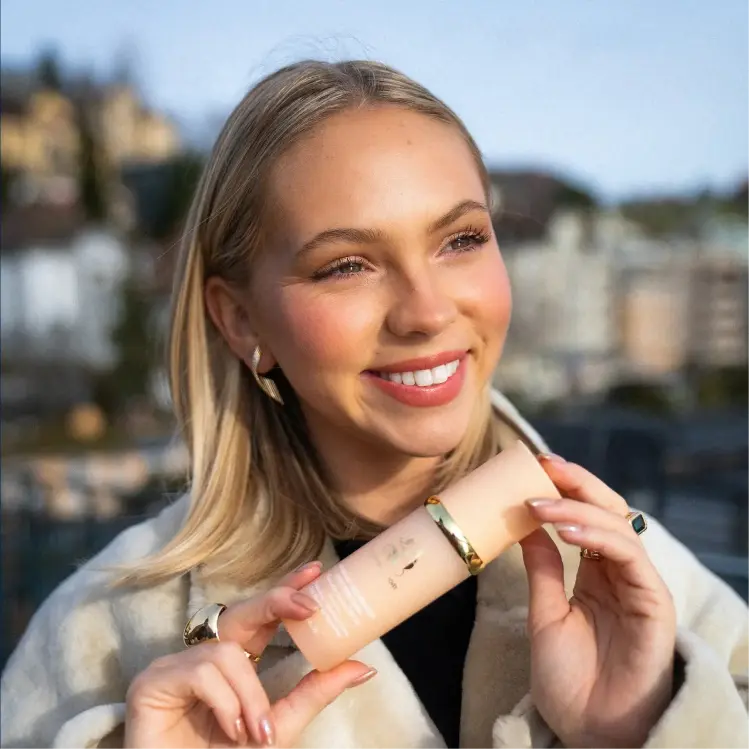 A smiling blonde woman in a white coat holds a pink cosmetic bottle outdoors.