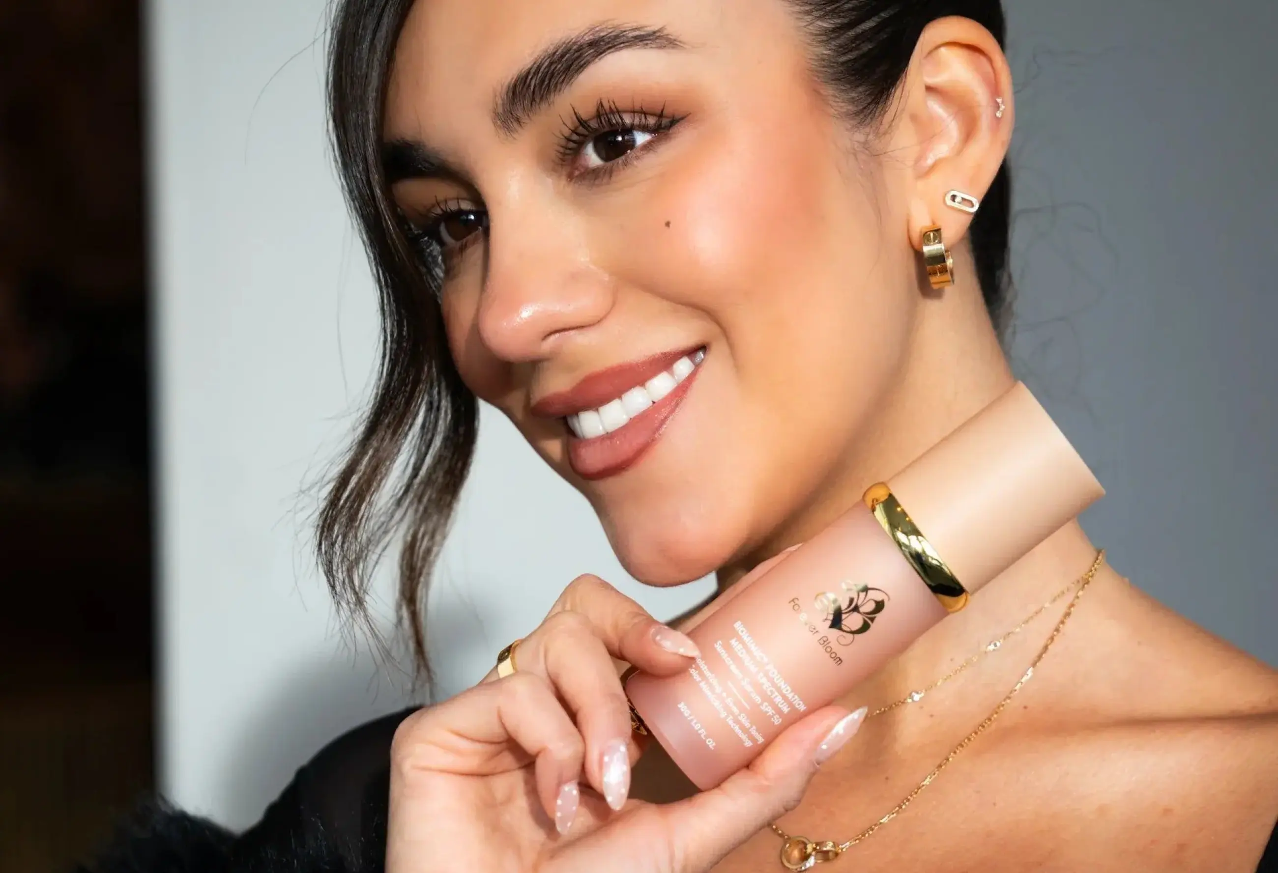 A smiling woman with dark hair and makeup holds a pink bottle of foundation.