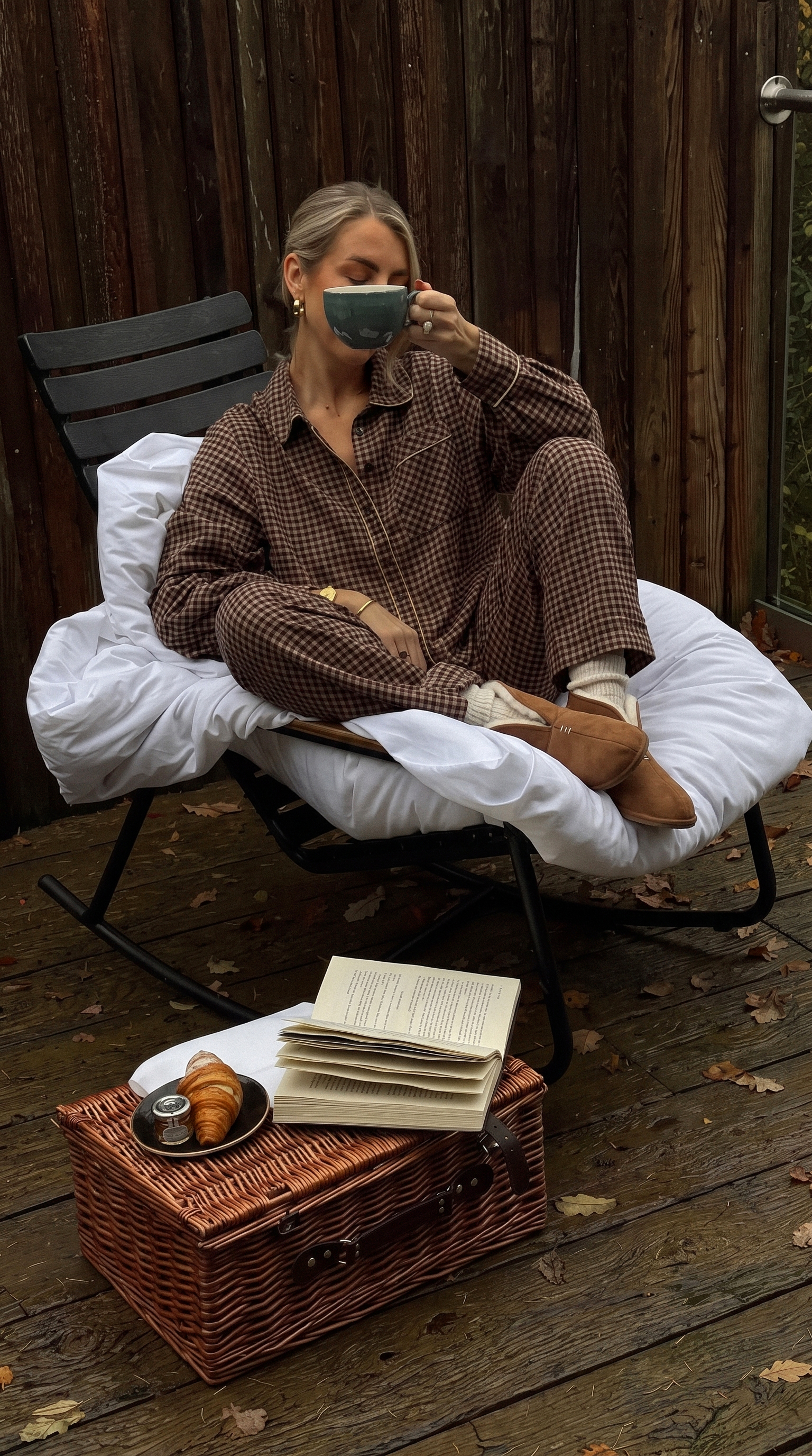 A woman in brown plaid pajamas sips a drink while sitting on a cushioned chair on a wooden deck.