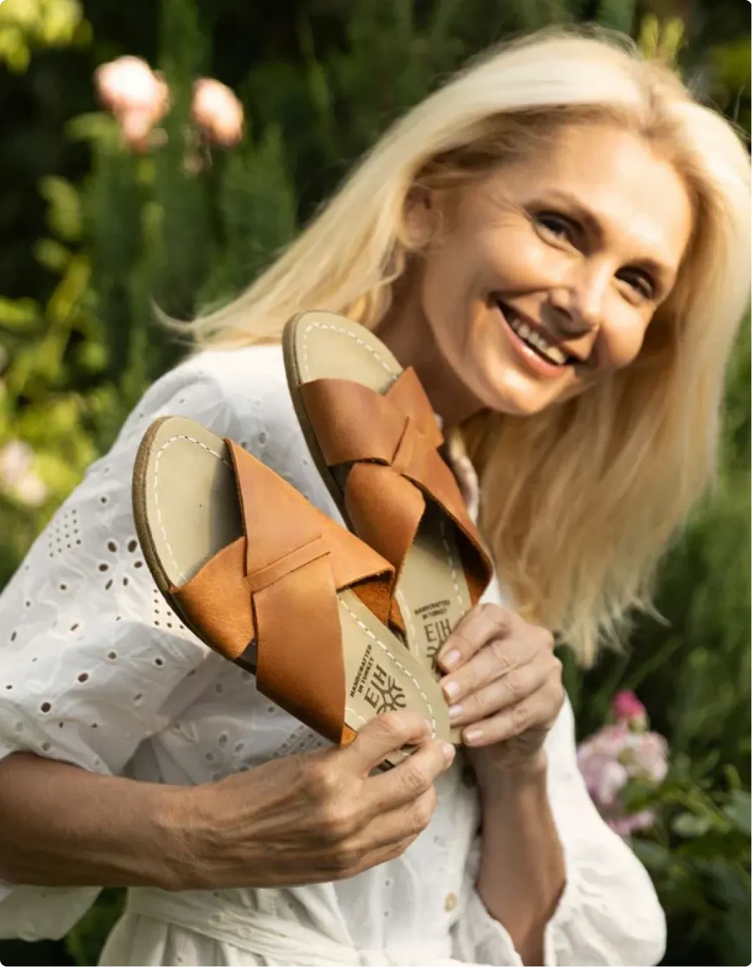 A smiling blonde woman in a white eyelet top holds up a pair of tan leather sandals.