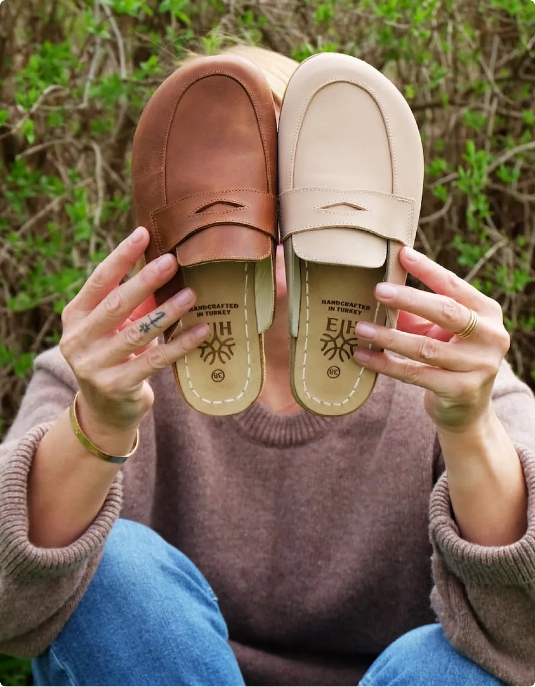 A person sits outdoors holding two different colored leather loafers in front of their face.