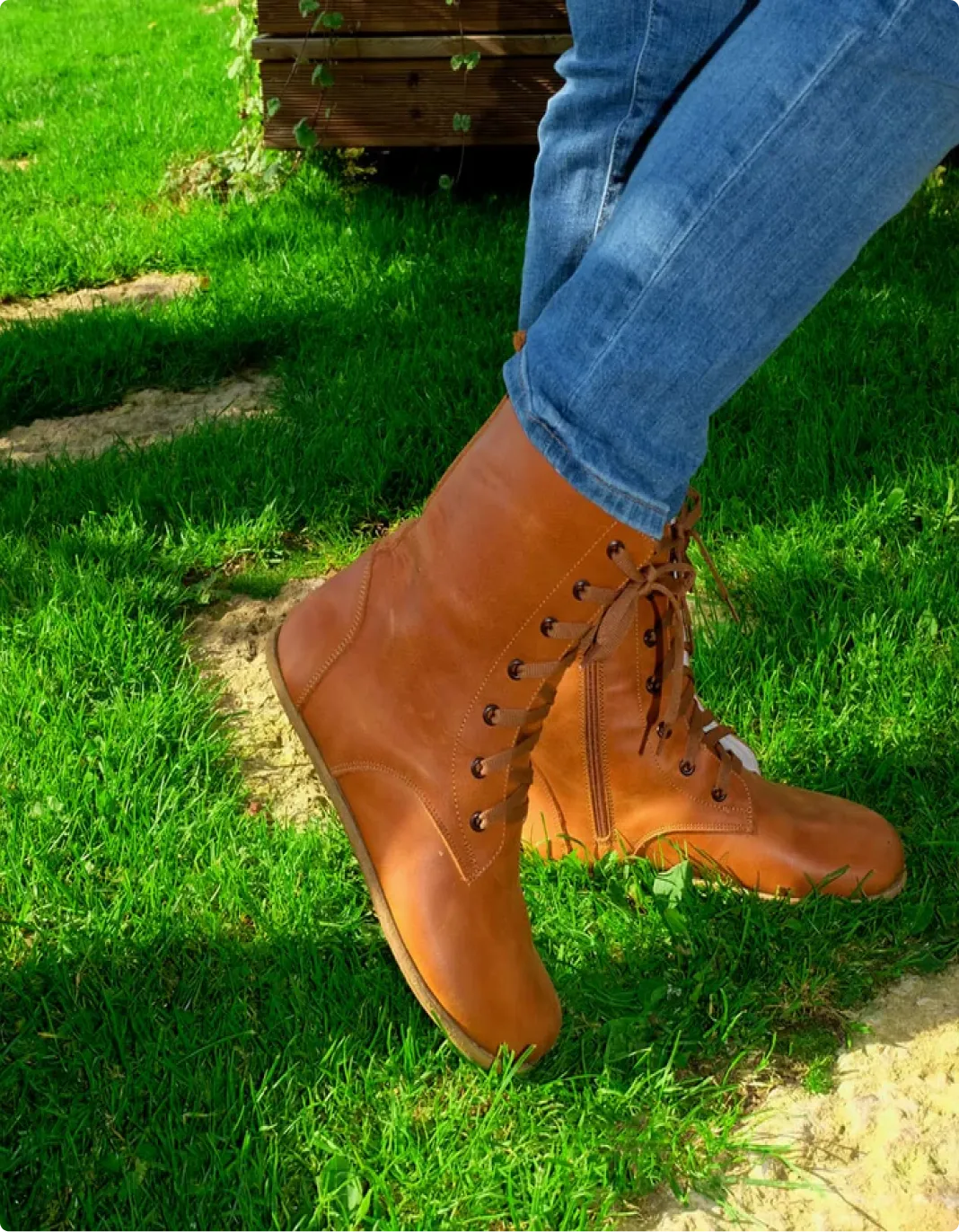 A person wearing blue jeans and brown lace-up boots rests their feet on a patch of grass.