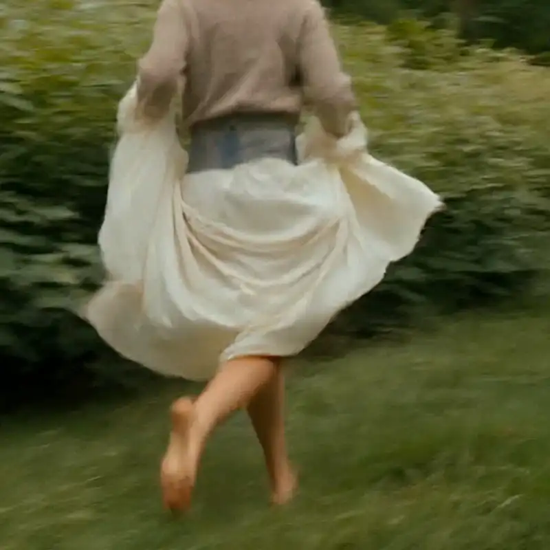 The back of a person wearing a flowing white skirt running barefoot through a grassy field.