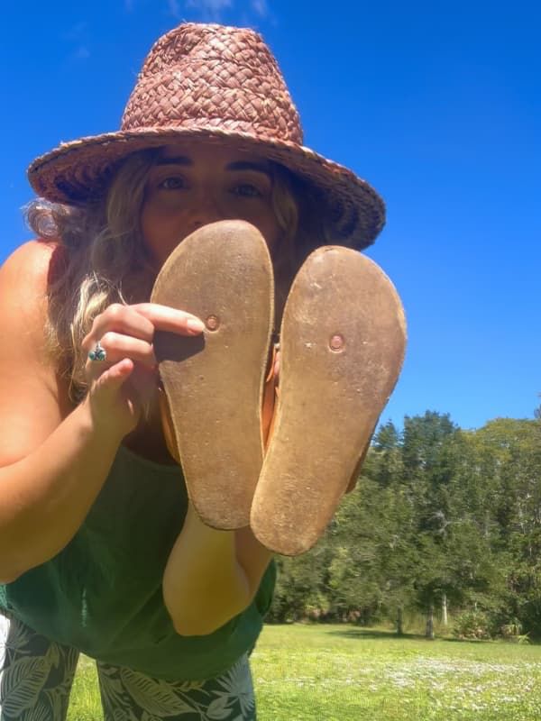 A person in a straw hat holds up the soles of their sandals to the camera outdoors.