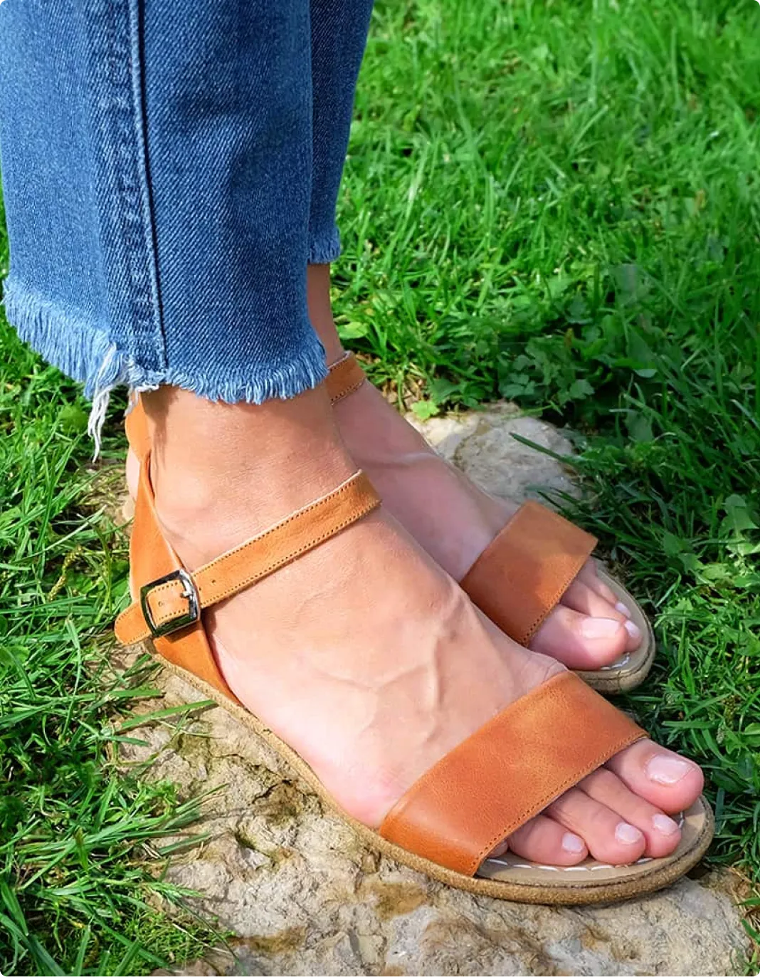 A person wearing frayed-hem jeans and brown leather sandals stands on a rock in the grass.