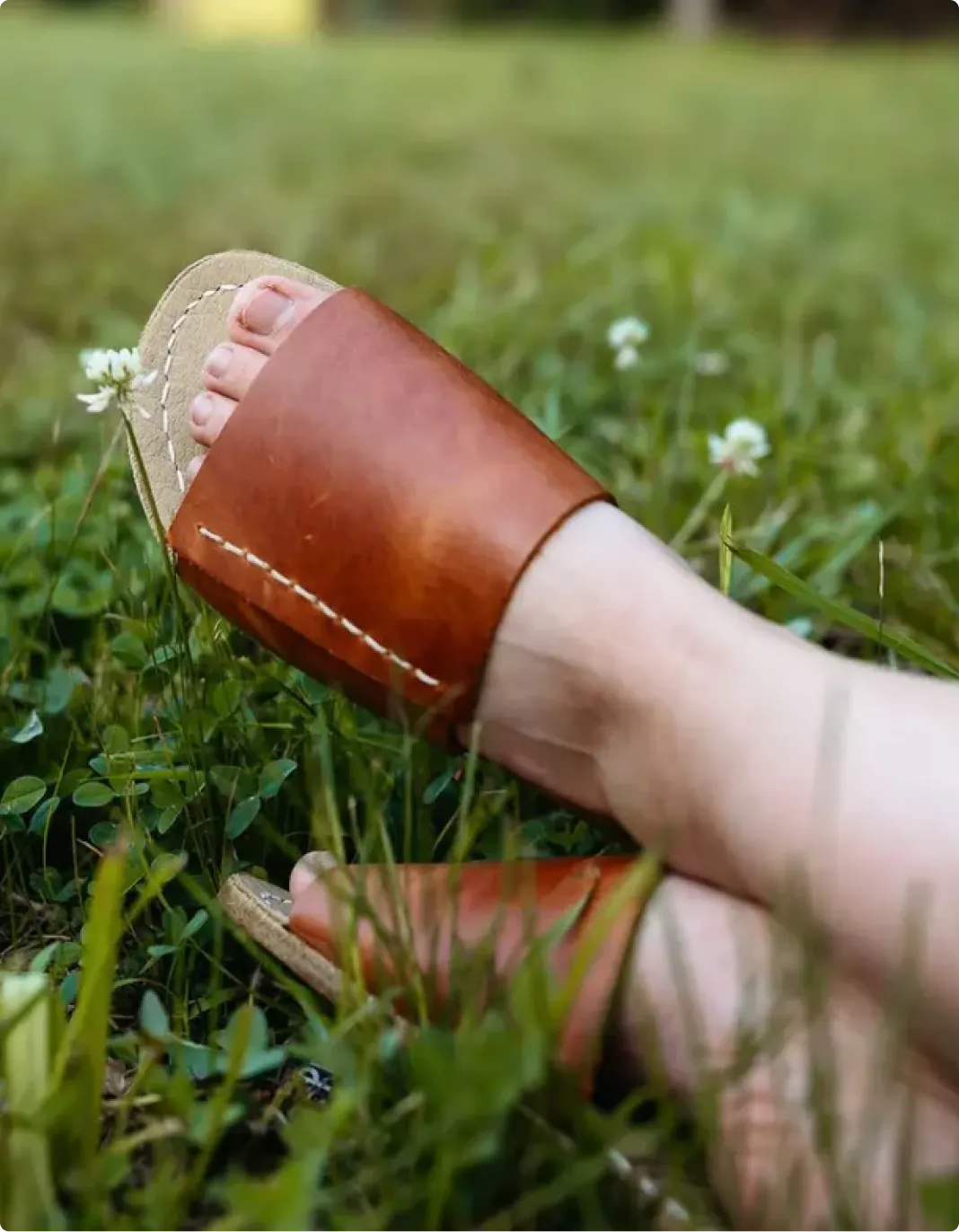 A person's feet in brown leather slide sandals with white stitching, resting in green grass.