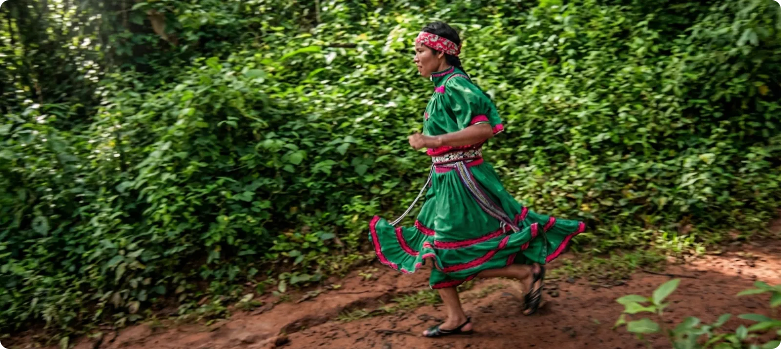 A person in a traditional green and pink dress runs on a dirt path in a lush forest.