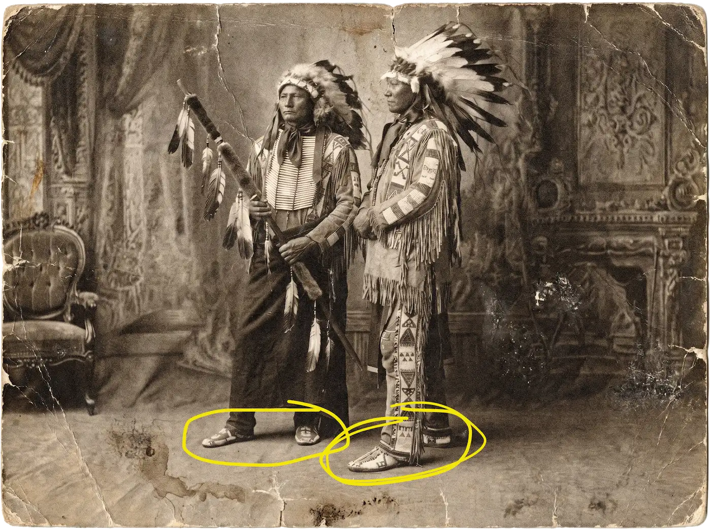 A vintage, damaged sepia photograph of two Native American men in traditional regalia and feathered headdresses.