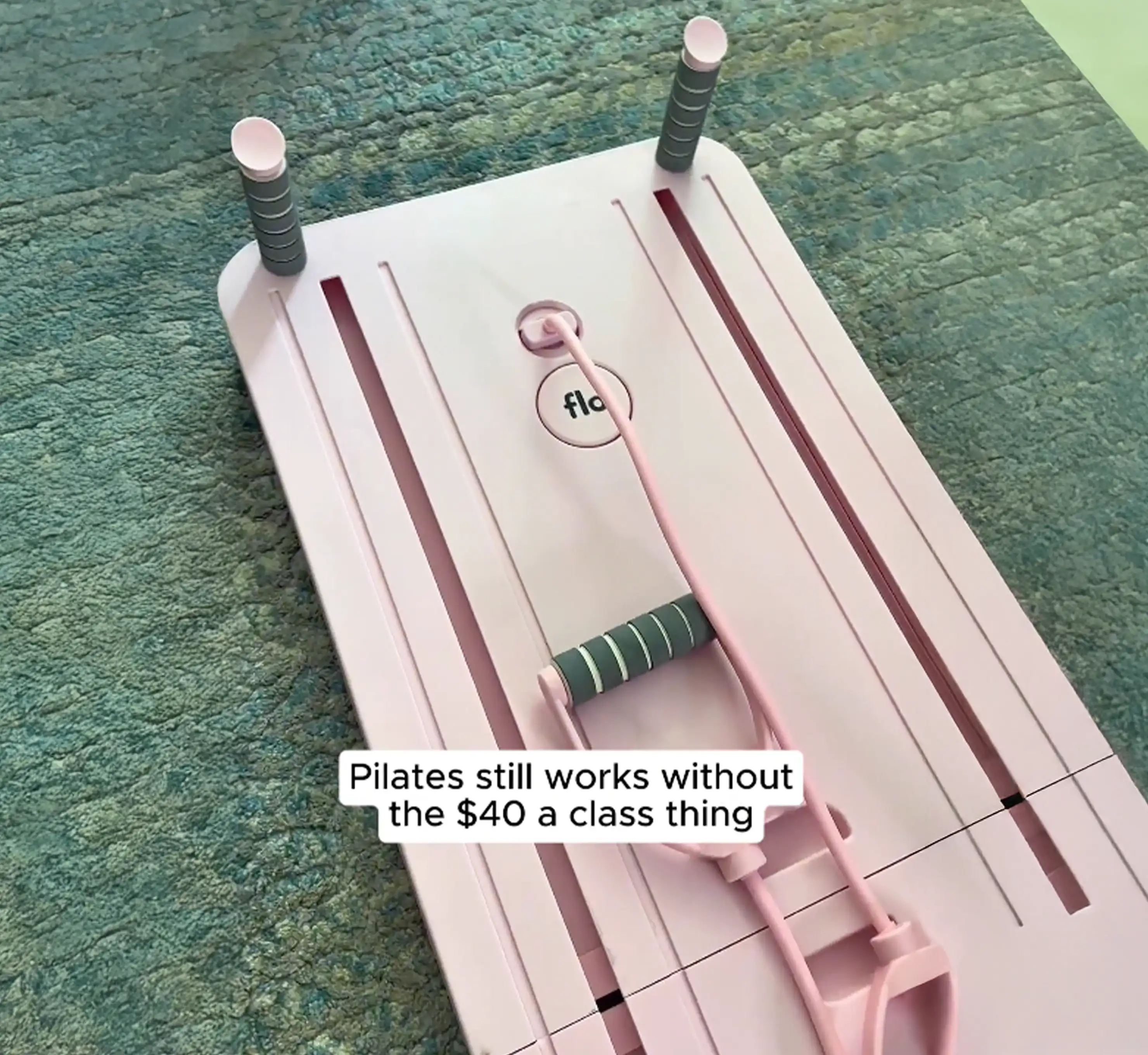 A pink portable pilates reformer with resistance bands lies on a teal rug.