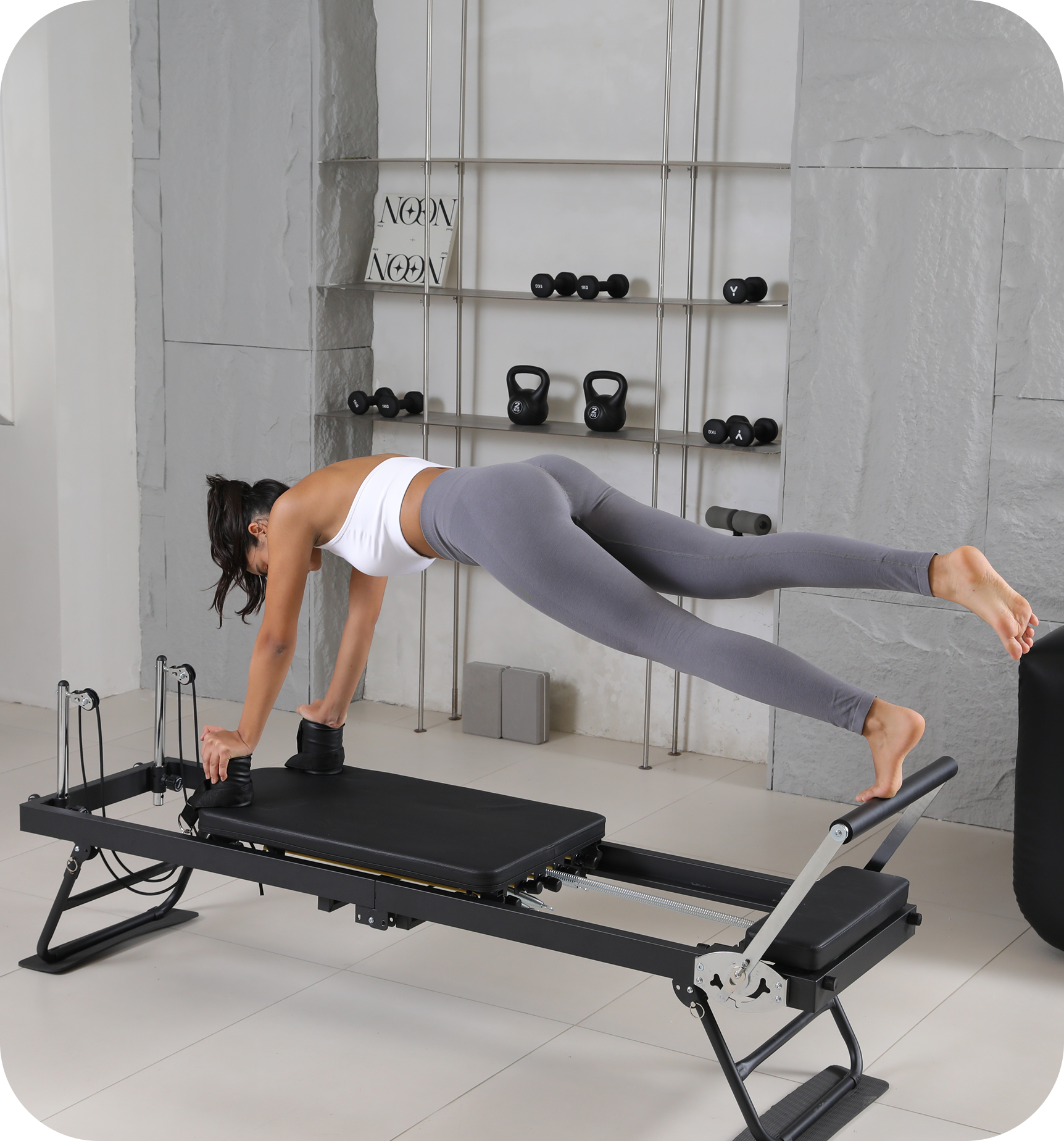 A woman in grey leggings and a white top performs an exercise on a Pilates reformer machine.