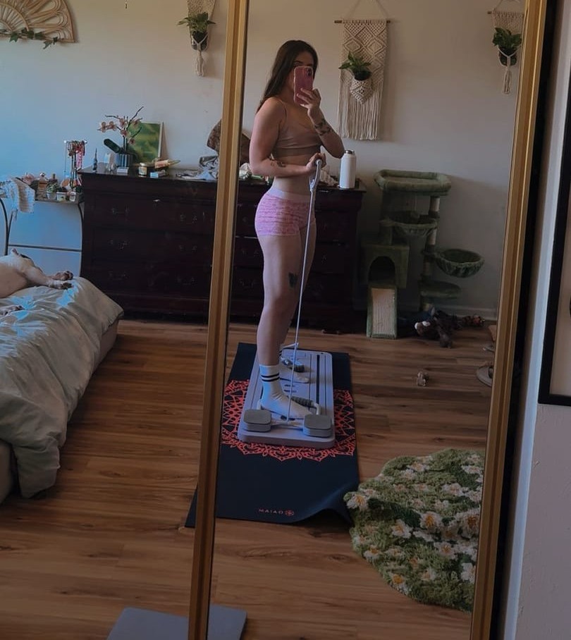 A woman in workout clothes takes a mirror selfie while standing on exercise equipment in a bedroom.