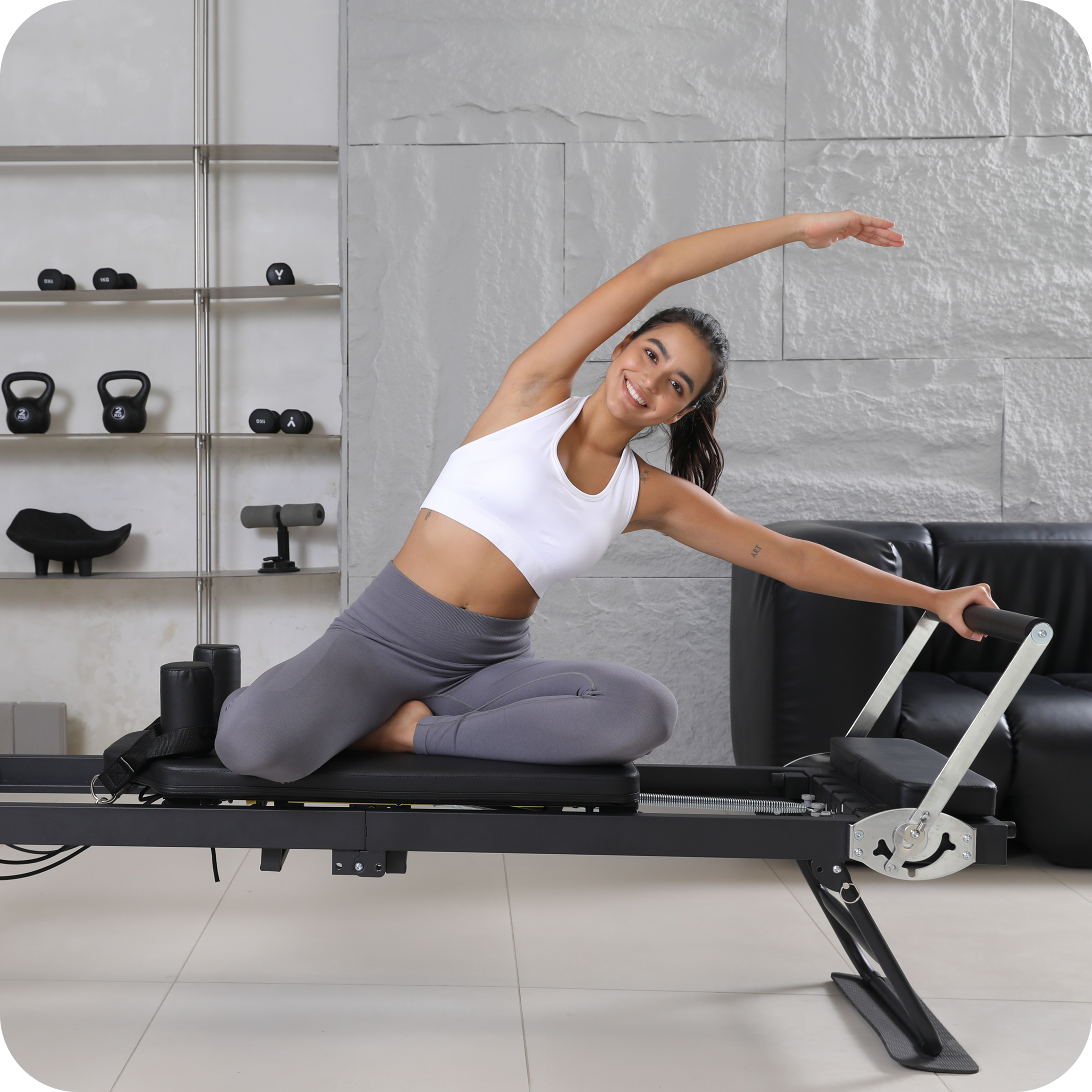 A smiling woman in sportswear stretching on a Pilates reformer machine in a home gym.