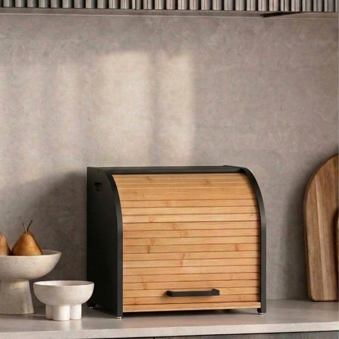 A black and bamboo roll-top bread box on a kitchen counter next to bowls and a cutting board.