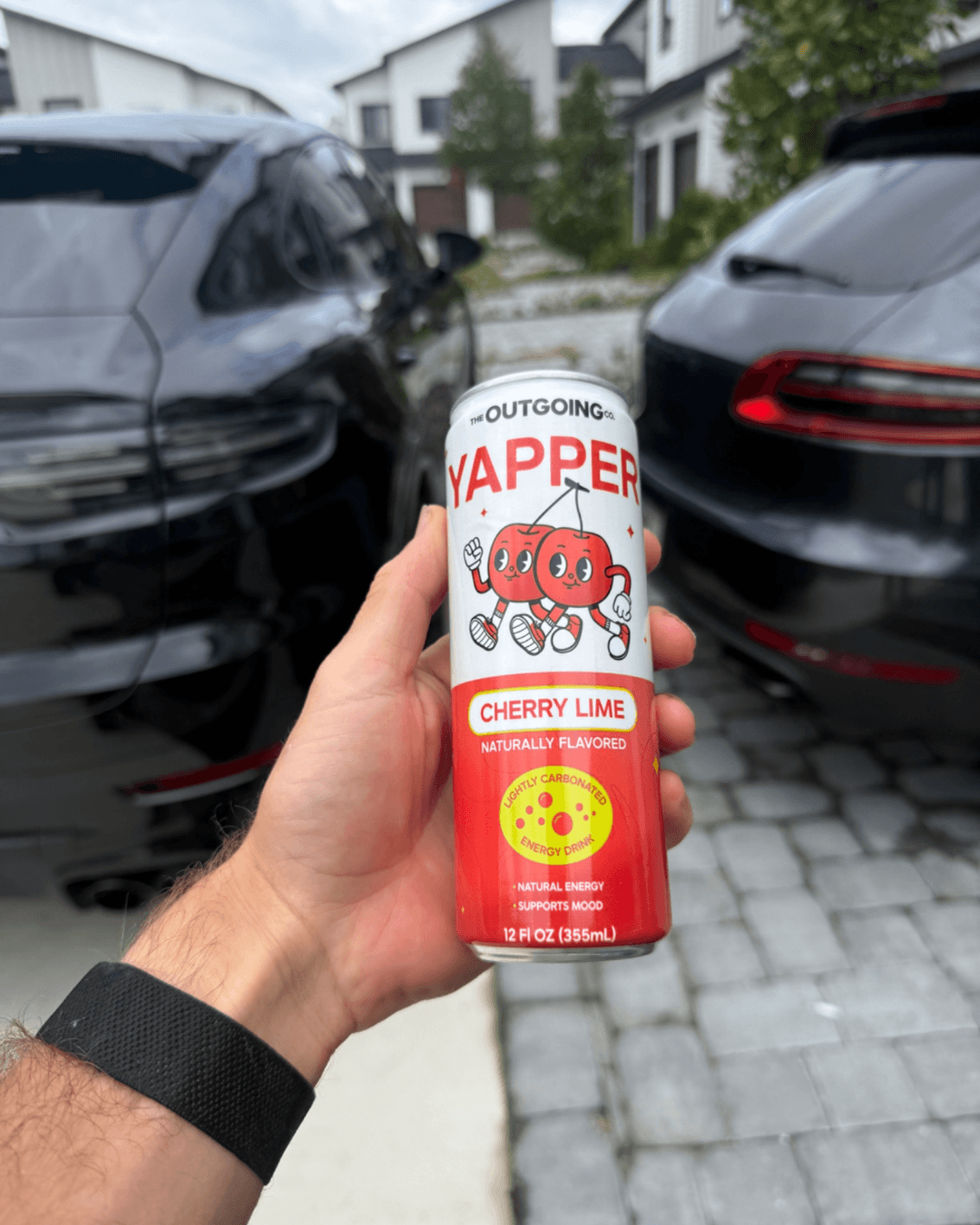 A hand holds a can of Yapper Cherry Lime energy drink in a driveway with two black cars.
