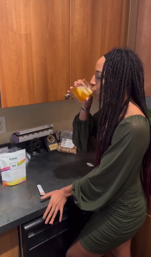 Person drinking orange juice in a kitchen, with supplements on the counter.