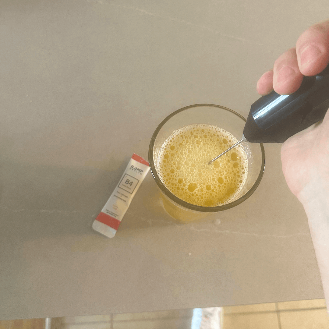 Hand frothing a yellow drink in a glass with a tool beside a sachet.